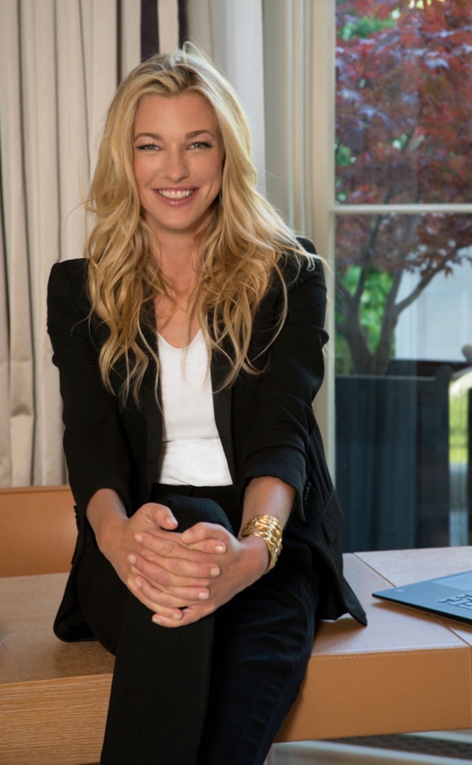 Portrait of Nicole Junkermann smiling warmly, seated on a desk beside a bright window