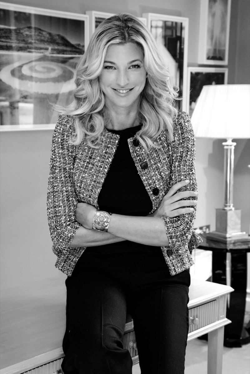 Black-and-white portrait of Nicole Junkermann in a tweed jacket, arms crossed, seated on a desk